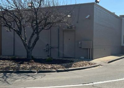 A leafless tree stands by the drive-thru lane of a gray building under a clear blue sky.