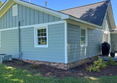 A small light blue house with white trim, brick foundation, and a deck with a grill on a sunny day.
