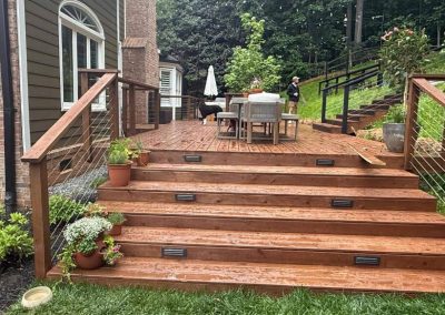 Wooden deck with stairs, potted plants, outdoor dining table and chairs, and a woman standing in the background.