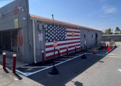 Long gray building with large American flag mural, red poles, cones, and accessible ramp in parking lot.