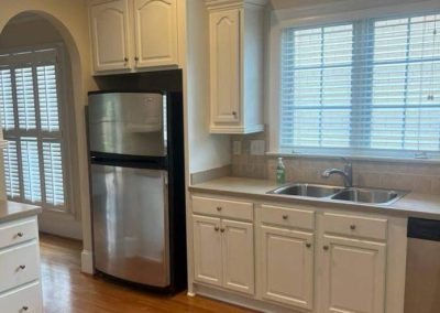 Kitchen with white cabinets, stainless steel fridge, double sink, window with blinds, and hardwood floor.