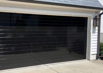 A modern, shiny black garage door on a white house with gray roof shingles and a concrete driveway.