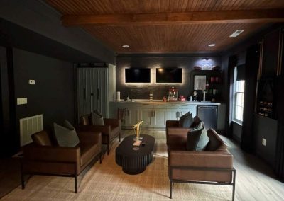 A modern lounge with brown chairs, a round table, and a bar area with two TVs on the back wall.