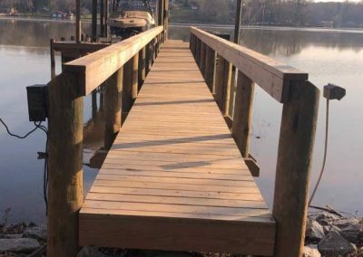 Wooden dock with railings extending over a calm lake toward a covered boat slip, trees in background.
