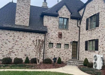 Two-story brick house with dark roof, trimmed shrubs, bare trees, and an HVAC unit on the right side.