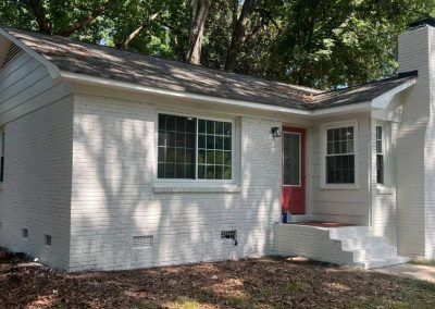 Small white brick house with a red door, large windows, and shady trees in the background.