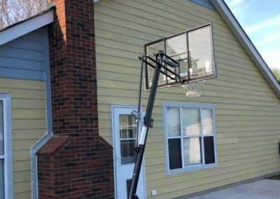 A basketball hoop is positioned in front of a yellow house with a brick chimney and windows.