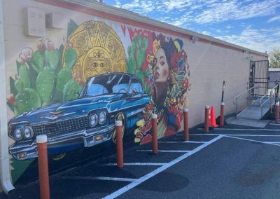 Mural of a classic car, a woman in traditional attire, and Aztec motifs on a building wall beside a parking lot.