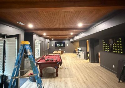 Large basement game room with pool table, ladder, and tennis-themed decor under wood-paneled ceiling.