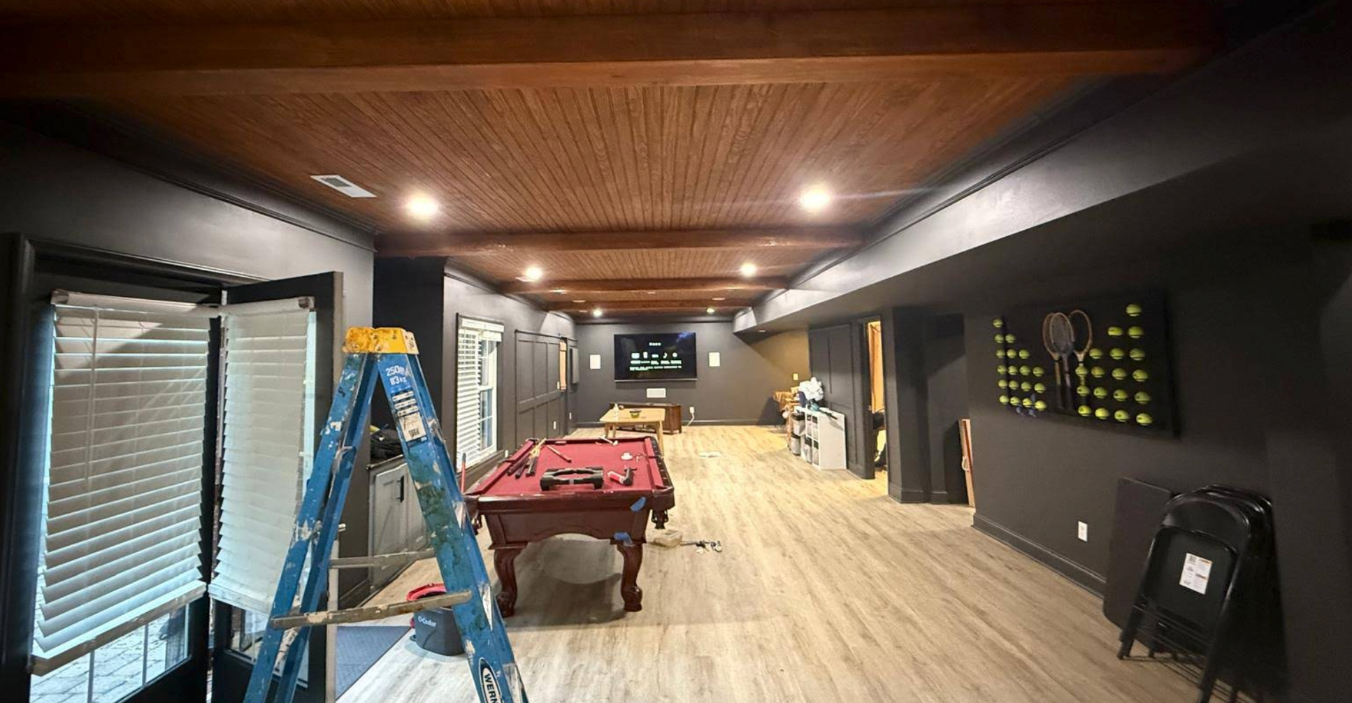 Large basement game room with pool table, ladder, and tennis-themed decor under wood-paneled ceiling.