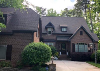 Brick house with steep roof, surrounded by trees and bushes, with a driveway and basketball hoop in front.