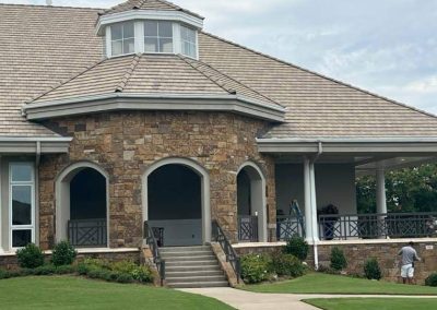 Large stone house with arched entry, stairs, covered porch, and workers painting near the side railing.
