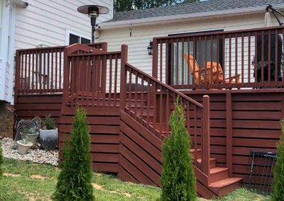 Red wooden deck with stairs, patio chairs, and tall shrubs in front of a white house.