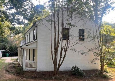White two-story house with a porch, surrounded by trees and grass on a sunny day.
