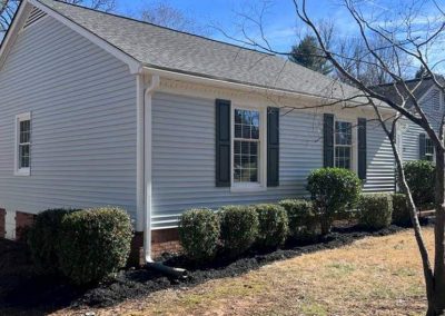Single-story house with light gray siding, dark shutters, and trimmed bushes in front, on a sunny day.