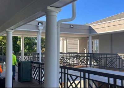 Two workers stand on a covered terrace with white columns and black railings under a clear blue sky.