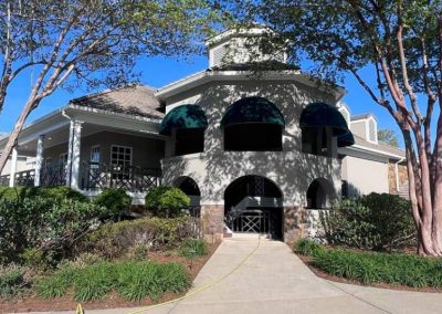 Two-story building with green awnings, surrounded by trees and bushes, under a clear blue sky.