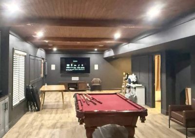 A game room with a pool table, TV, chairs, and various sports equipment under wooden ceiling beams.