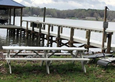 White picnic table near a lakeside dock with a boat, surrounded by grass, chairs, and overcast sky.