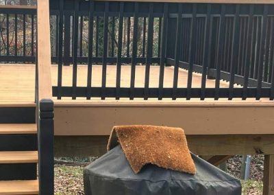 A Weber grill covered with fabric sits below a wooden deck with black railings and stairs.