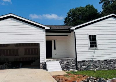 A white house with stone trim, an open garage, and unfinished landscaping under a blue sky.