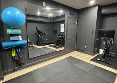A home gym with exercise equipment, wall mirror, yoga mats, and a stationary bike on wood and mat flooring.