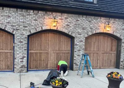 Person kneeling in front of a wooden garage door; ladder and potted flowers are nearby on a driveway.