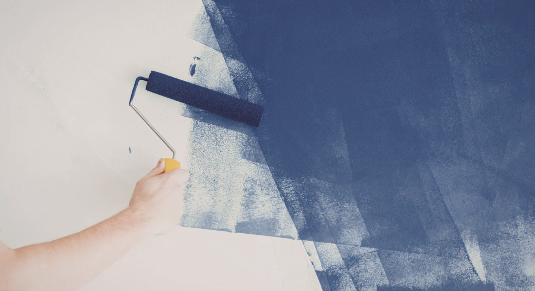 A hand uses a paint roller to apply dark blue paint to a white wall.