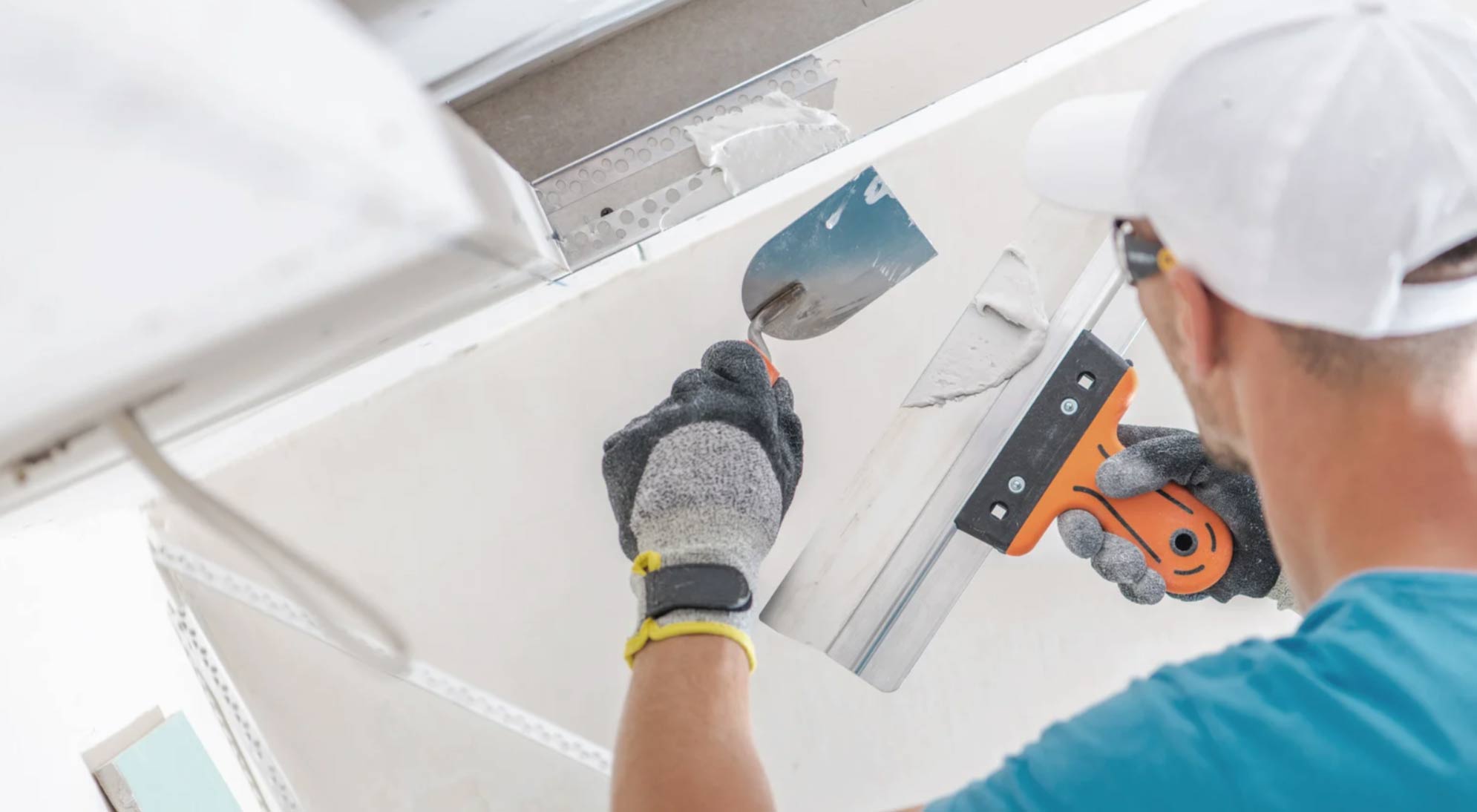 Person wearing gloves and a cap using tools to apply plaster or finish a white ceiling.