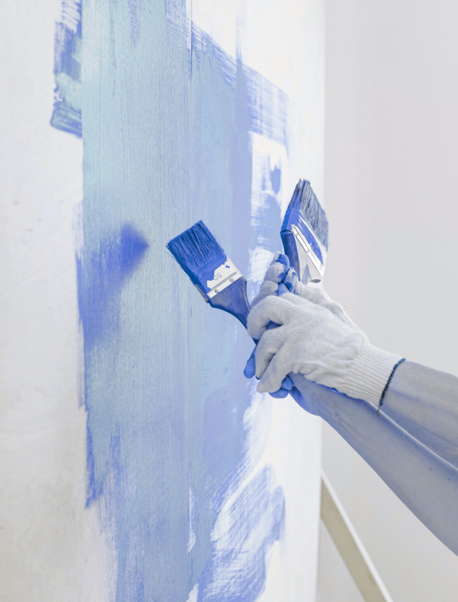 A person wearing gloves paints a wall light blue with two wide brushes.