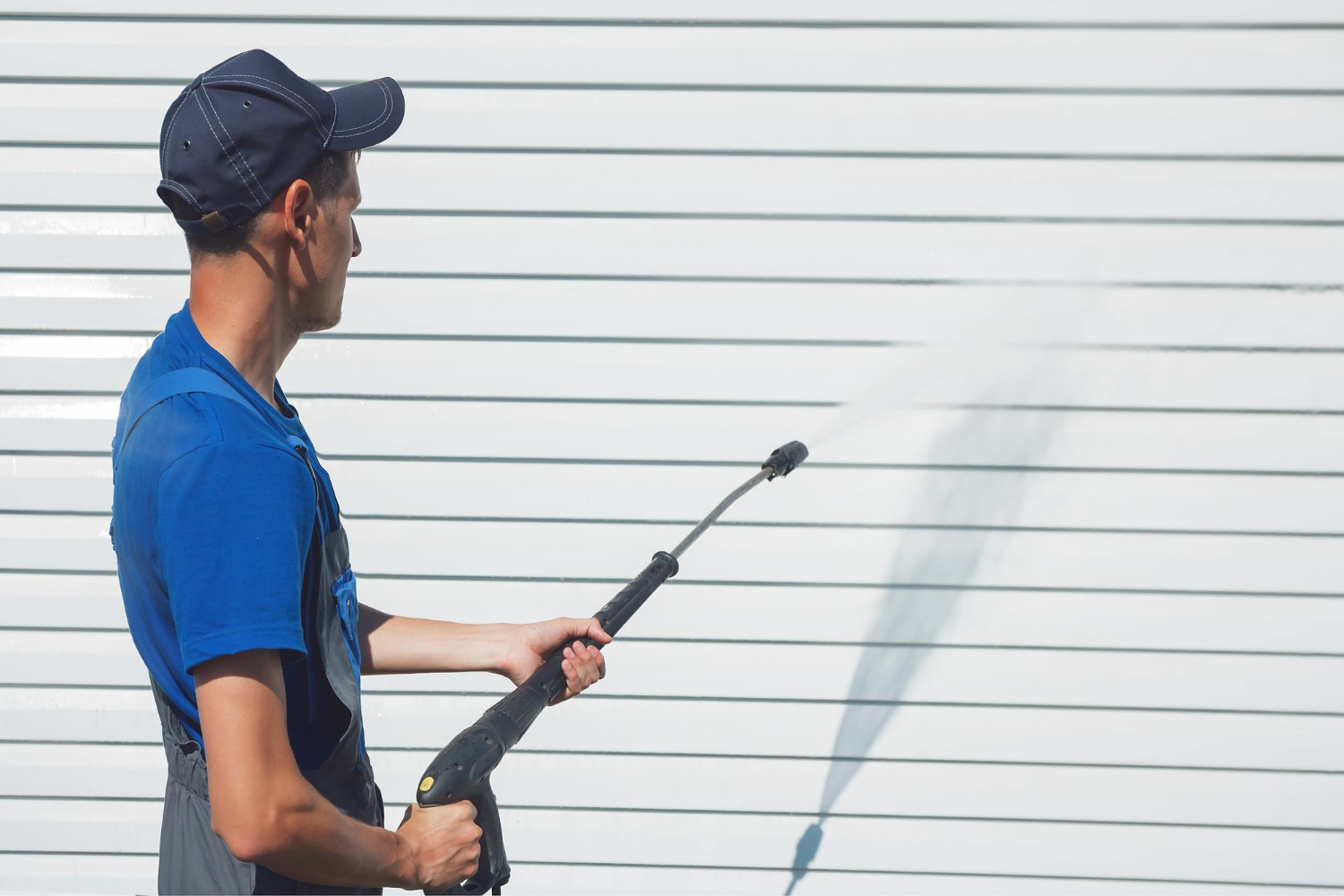 Man in blue shirt pressure washing a white garage door, water spraying from the nozzle.